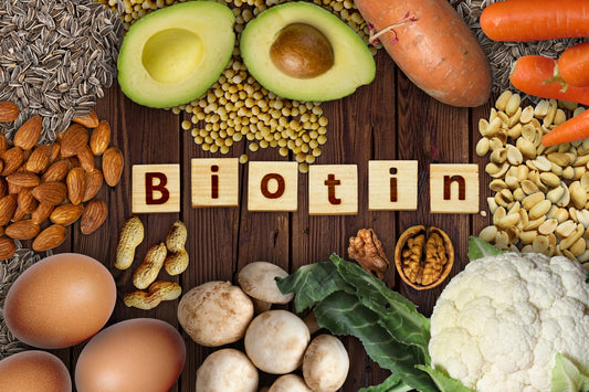 Food sources of biotin on a table, including eggs, almonds, peanuts, avocado, sunflower seeds, and a 'Biotin' sign in blocks.