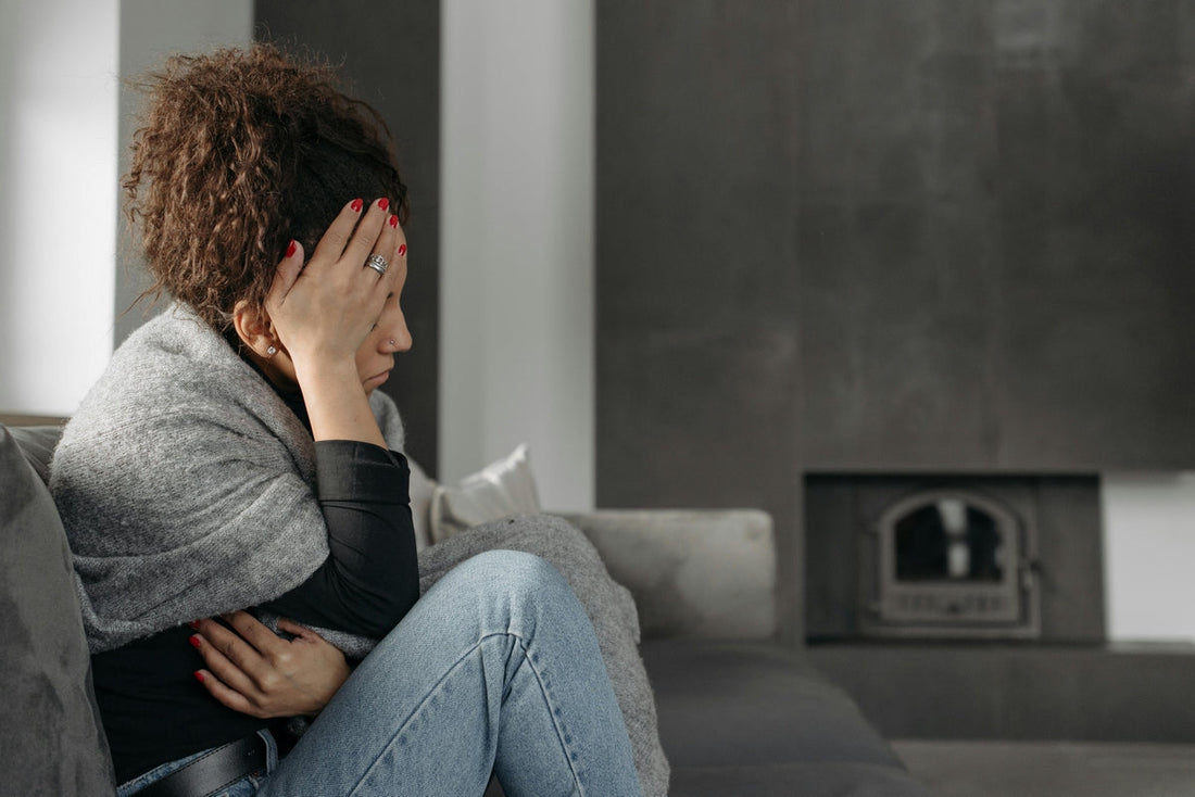 A woman sits on a gray sofa, wrapped in a blanket, with her head resting on her hand in a pensive or sad pose