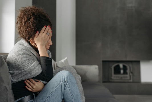 A woman sits on a gray sofa, wrapped in a blanket, with her head resting on her hand in a pensive or sad pose